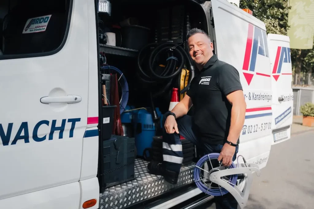 Ein Mitarbeiter von ARDO Abflussreinigung Düster & Ostermann OHG steht vor einer geöffneten Fahrzeugtür und lächelt in die Kamera